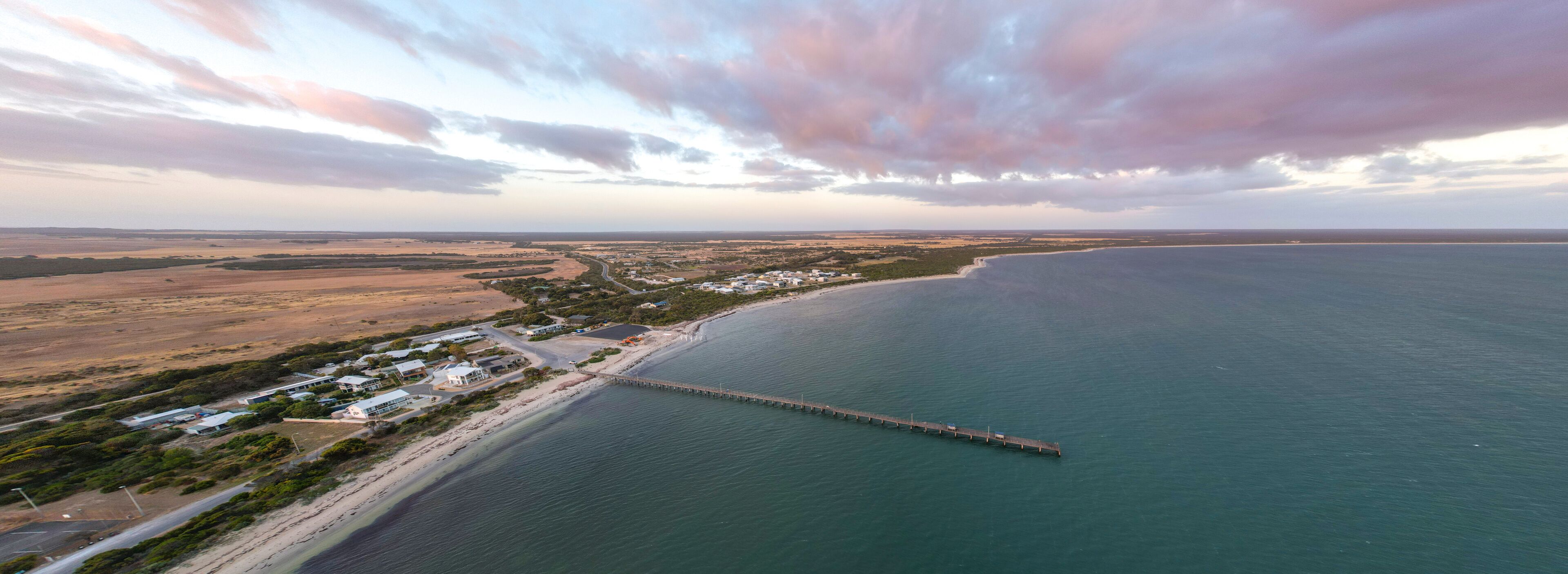 An aerial view of a coastal area with a long pier extending into the sea, surrounded by a small settlement with houses and roads.