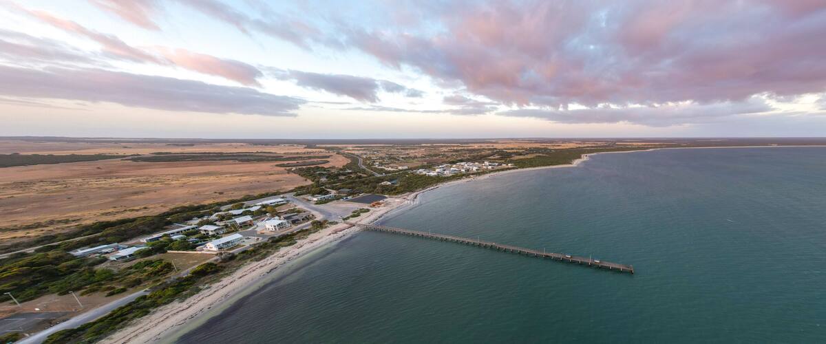An aerial view of a coastal area with a long pier extending into the sea, surrounded by a small settlement with houses and roads.