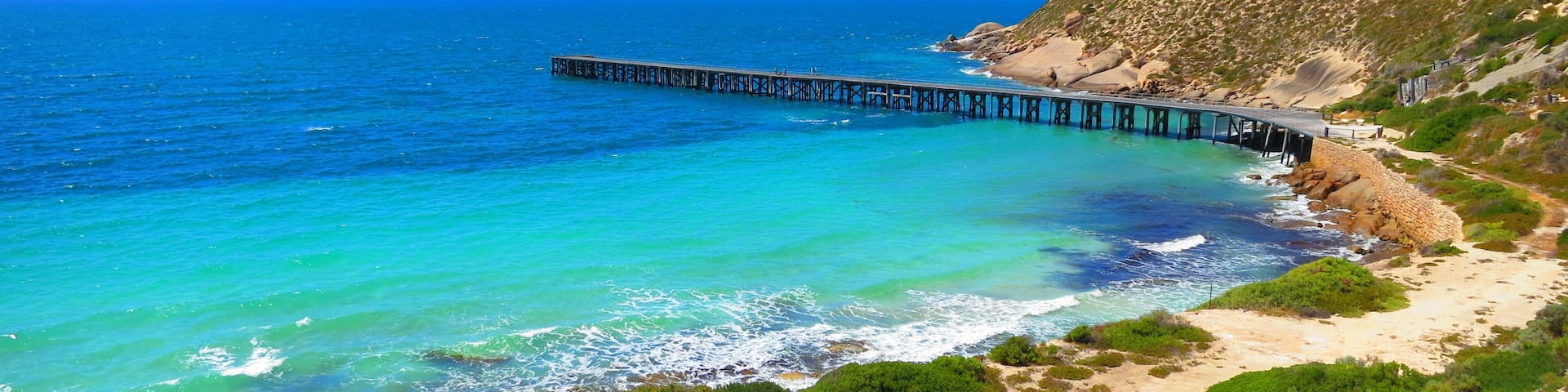 A coastal landscape with a pier extending into the turquoise sea, surrounded by green vegetation and rocky cliffs.