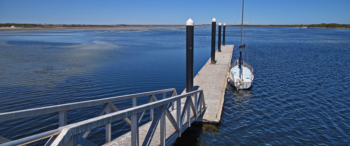 A calm lake with a dock extending into the water, a sailboat moored at the dock, and a clear blue sky.