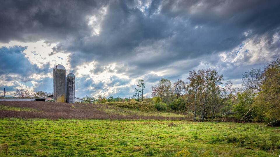 A rural landscape with a barn, silo, and trees under a cloudy sky.