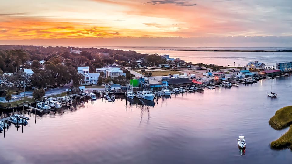 A marina with boats docked, a pier extending into the water, and a sunset over a coastal town.