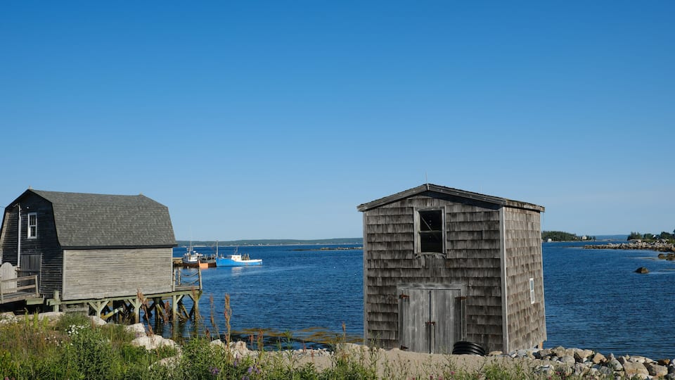 A wooden boathouse and a shed by a calm body of water with a boat in the background.