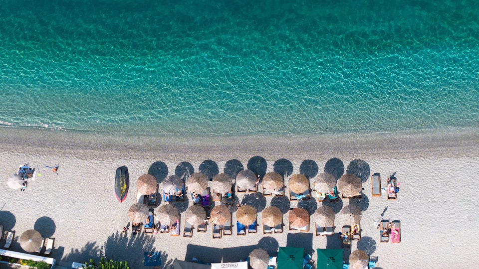 An aerial view of a beach with clear turquoise water, rows of sun loungers, and thatched umbrellas.