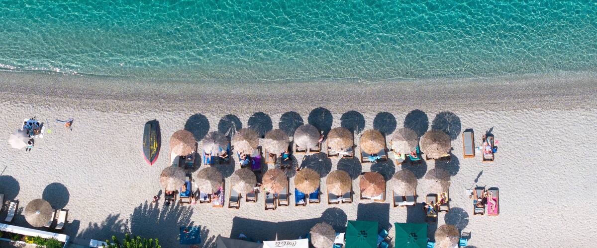 An aerial view of a beach with clear turquoise water, rows of sun loungers, and thatched umbrellas.