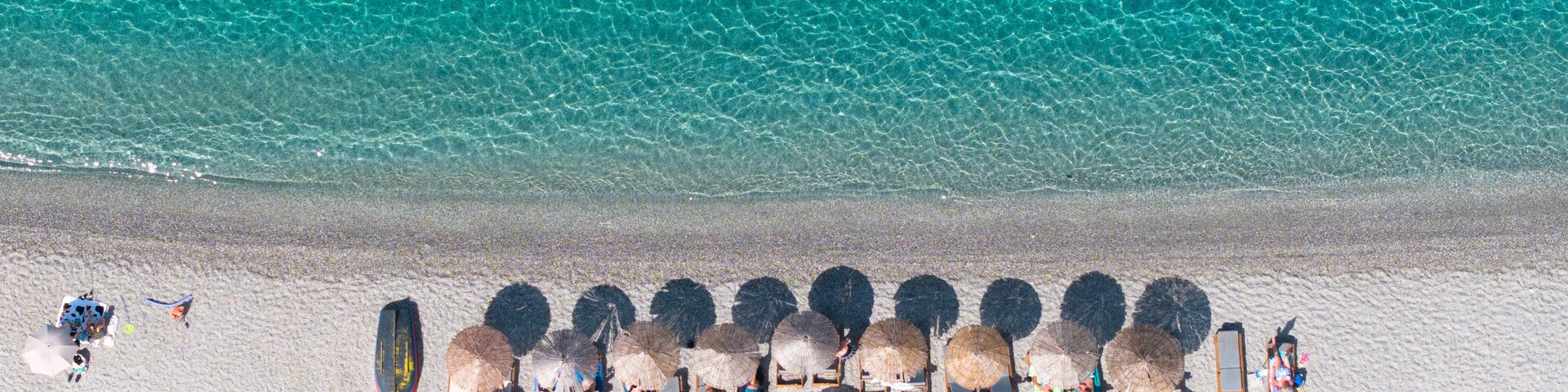 An aerial view of a beach with clear turquoise water, rows of sun loungers, and thatched umbrellas.