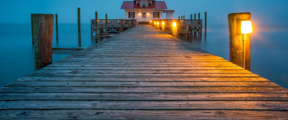 A wooden pier leading to a lighthouse with a red roof and a white building at the end.