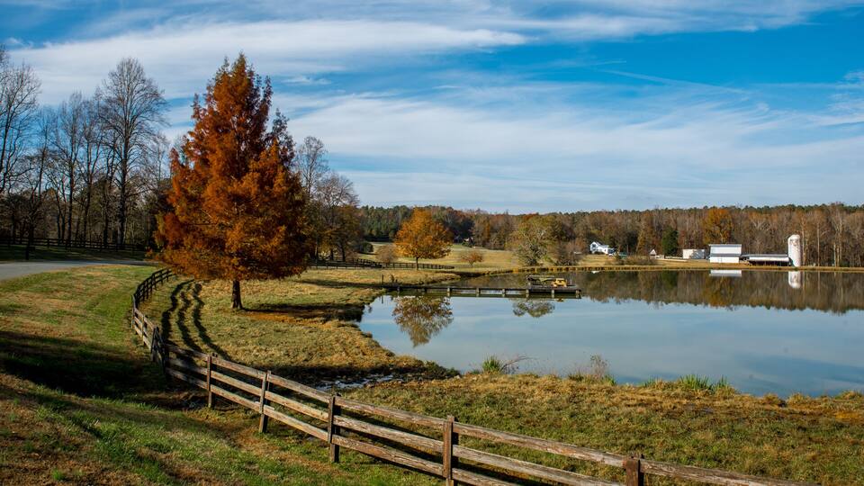 A serene lake with a wooden fence, autumn trees, and a barn in the background.