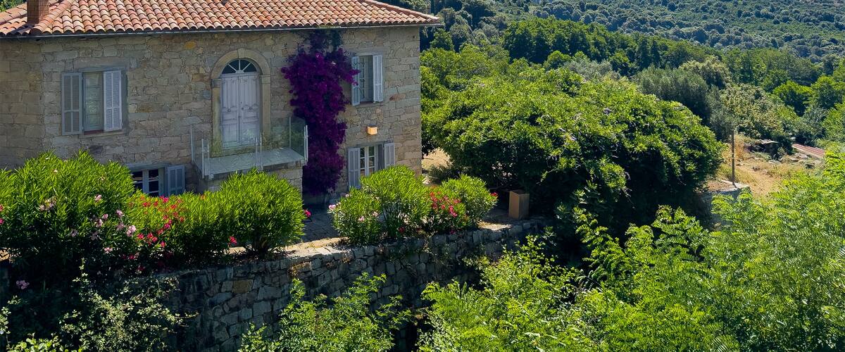 A stone house with a tiled roof surrounded by lush greenery and mountains in the background.