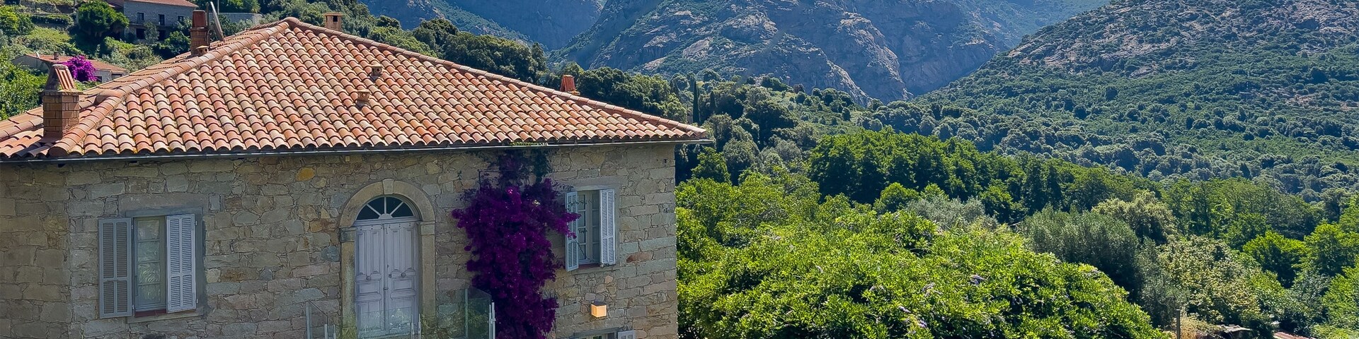 A stone house with a tiled roof surrounded by lush greenery and mountains in the background.