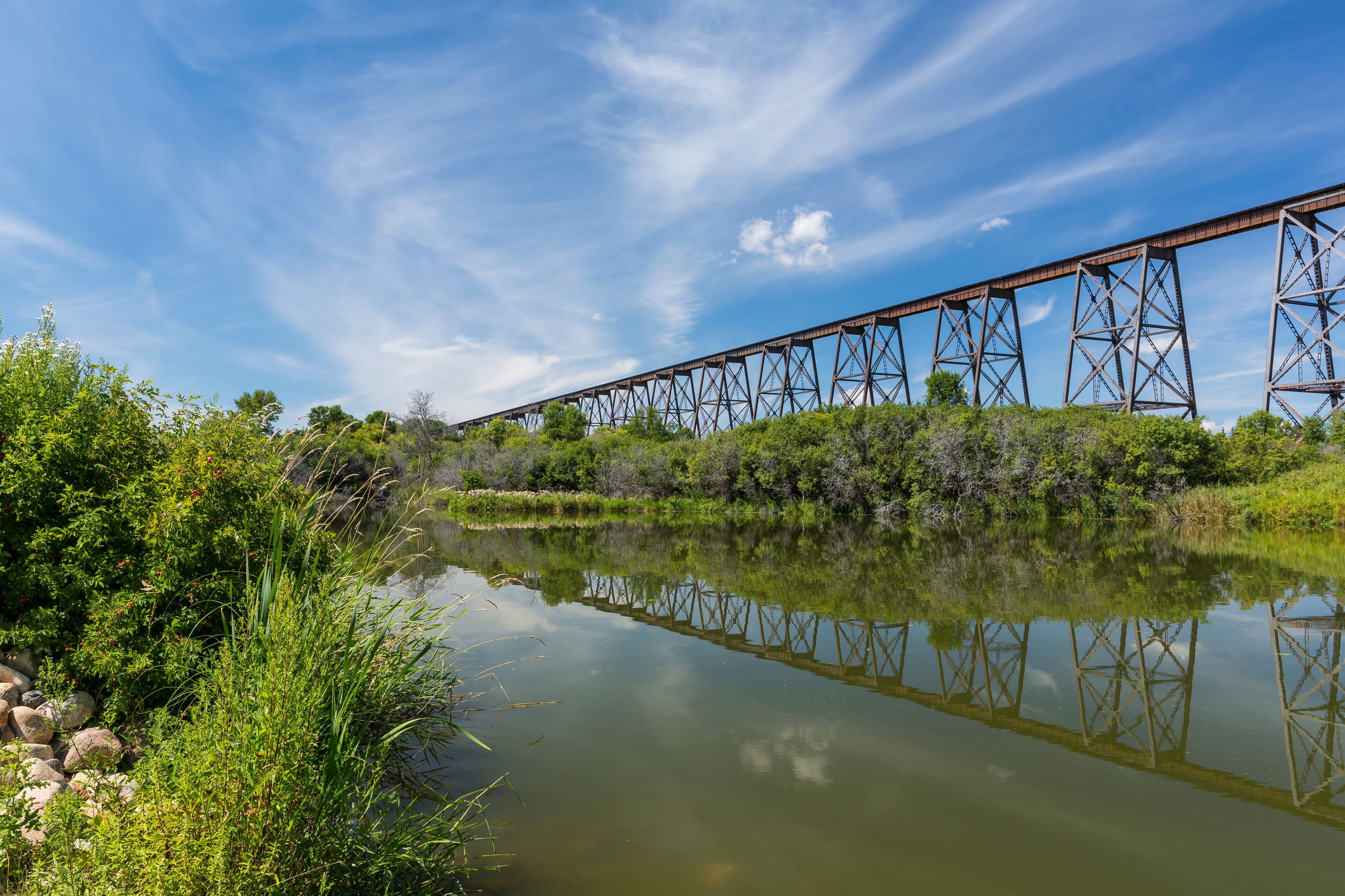 A steel bridge over a calm river, surrounded by lush greenery.