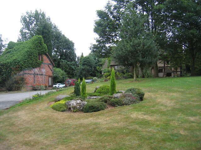 Grounds at Betteridge Farm. A shrubbery feature on the lawns leading down to Bakehouse Lane. The timber framed farm building can be seen through the trees.