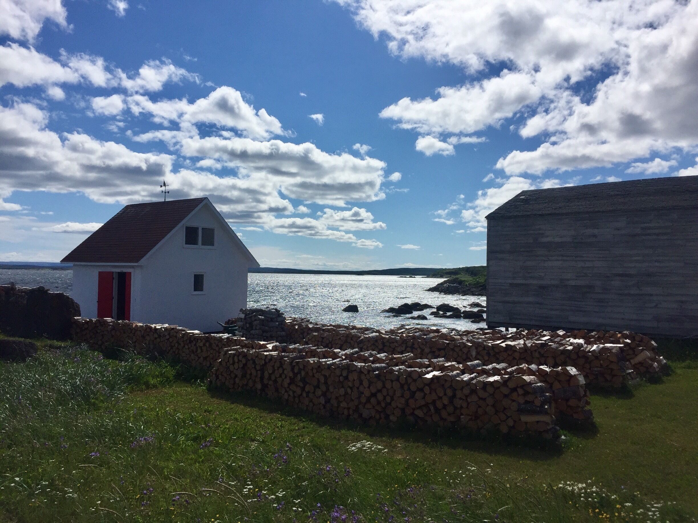 A white house with a red door, a stone wall, and a stone building by the water.