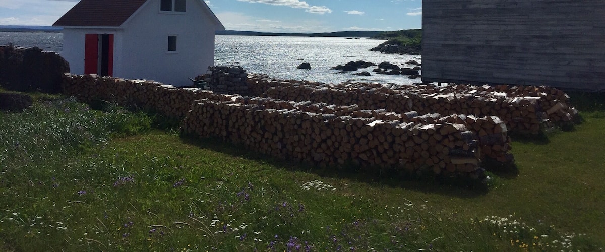 A white house with a red door, a stone wall, and a stone building by the water.