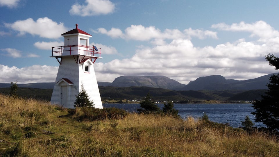 A white lighthouse with a red roof, surrounded by grass and trees, with mountains and a lake in the background.