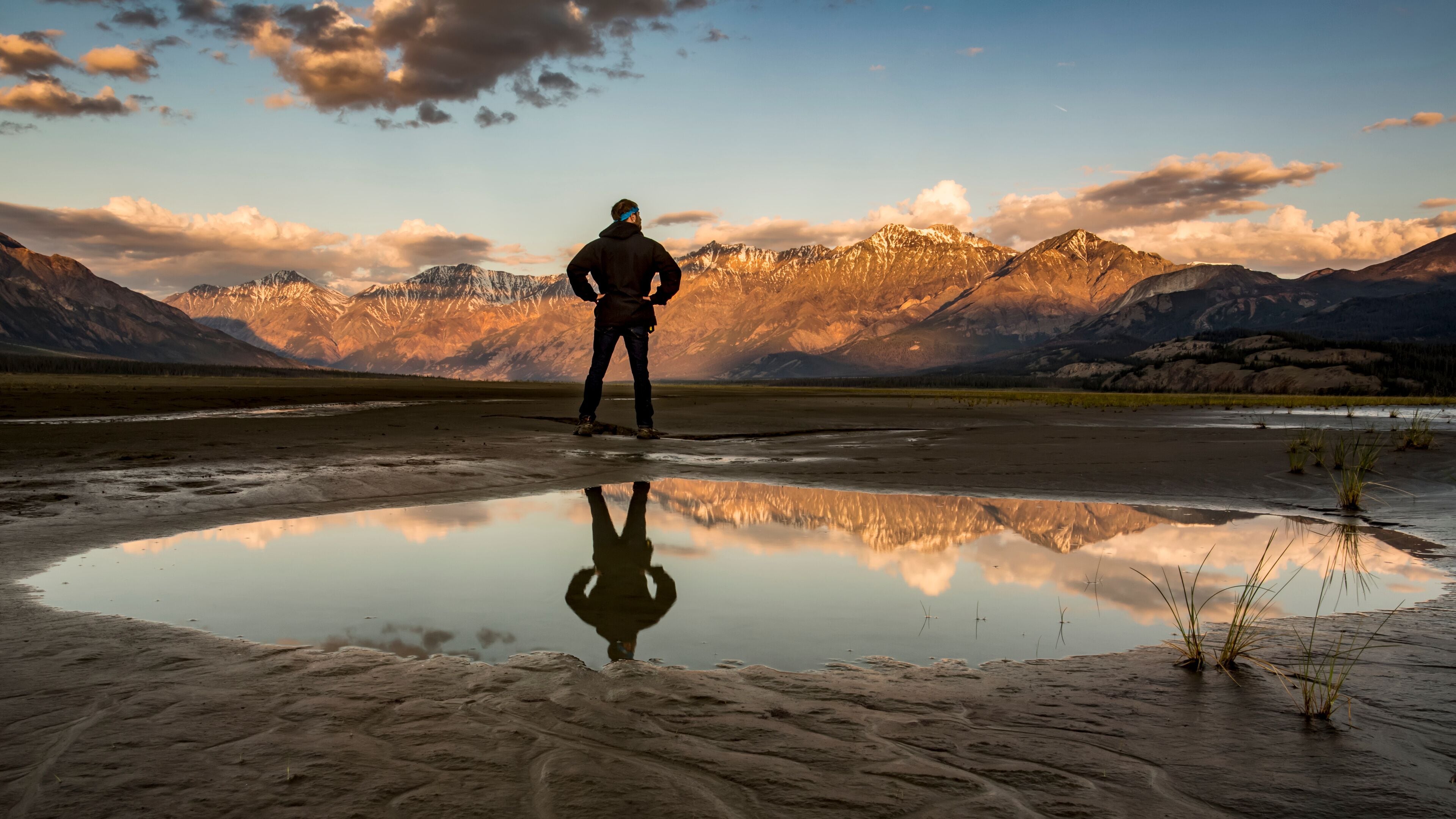 A man stands with his reflection in a pool of water looking out over the Saint Elias Mountains at sunset, Kluane National Park and Reserve; Destruction Bay, Yukon, Canada
