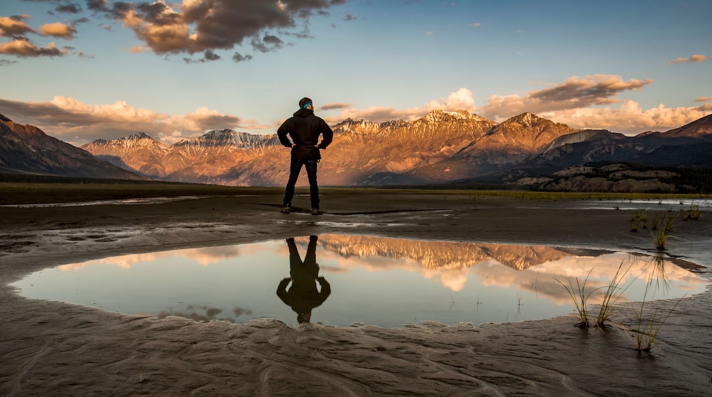 A man stands with his reflection in a pool of water looking out over the Saint Elias Mountains at sunset, Kluane National Park and Reserve; Destruction Bay, Yukon, Canada