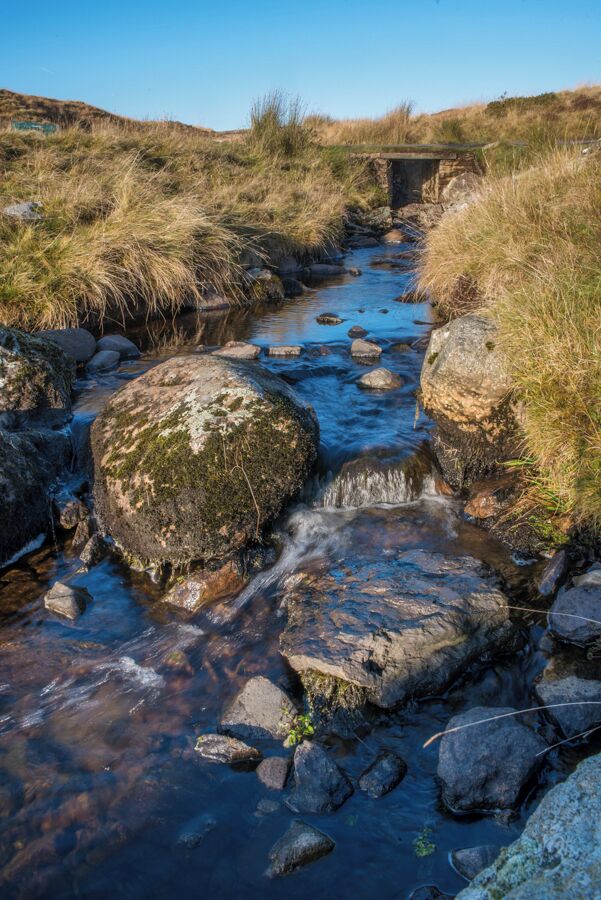 A stream flowing over rocks with a stone bridge in the background.