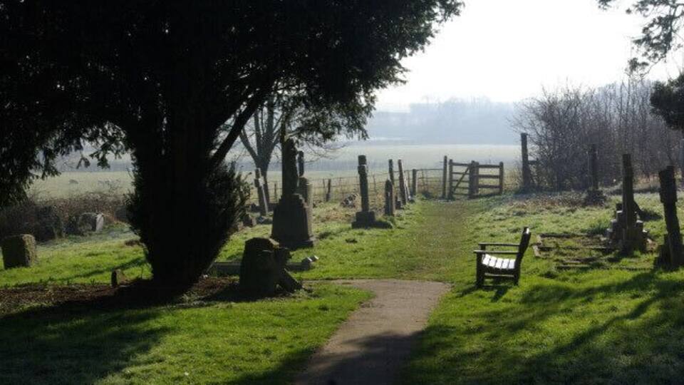 A cemetery with headstones, a bench, and a path leading to a distant view.