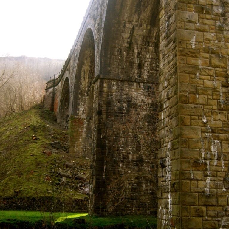 A stone bridge with multiple arches, surrounded by a grassy hillside and trees.