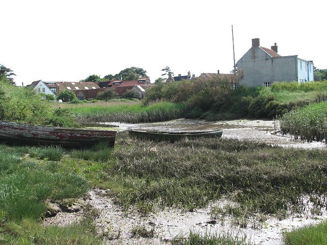 The tide is out The wet mud in the creek is glistening in the late afternoon sunshine; the view is southerly towards houses lining the Peddars Way & Norfolk Coast Path (seen in mid-distance). The house seen at right is located right at the edge of the saltmarsh. Many local families still make their living by fishing from Brancaster Staithe, which is famous for its first class shellfish. The harbour is also home to Brancaster Sailing Club, and sailing dinghies can be seen stored in and around the harbour. According to most books, Admiral Horatio Nelson learnt to sail at Brancaster but since in the mid to late 18th century there was no such thing as sailing for pleasure, it is more likely that he visited Brancaster Staithe from his home in nearby Burnham Thorpe, and persuaded local fishermen to take him out in their sailing boats.