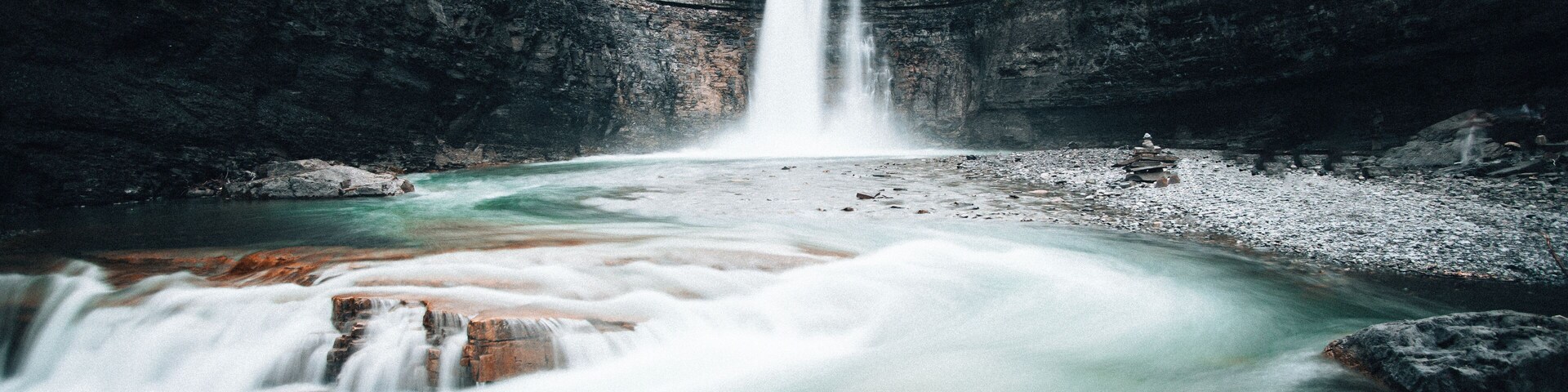 Una cascada que desemboca en un río, rodeado de acantilados rocosos y algunos árboles.