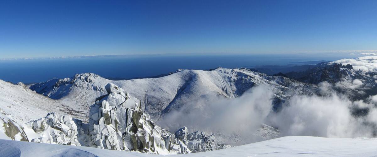 A snow-covered mountain range with a clear sky and a distant view of the ocean.