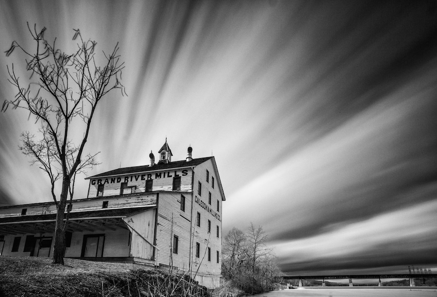 A historic building with 'Grand River Mills' signage, surrounded by bare trees and a cloudy sky.