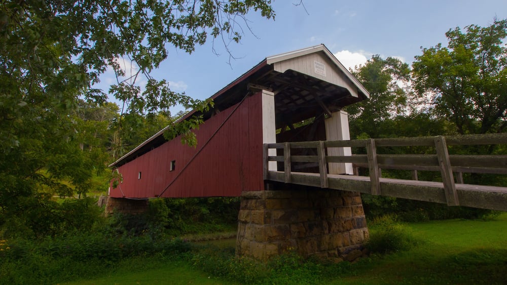 A red covered bridge with a stone foundation, surrounded by greenery and trees.