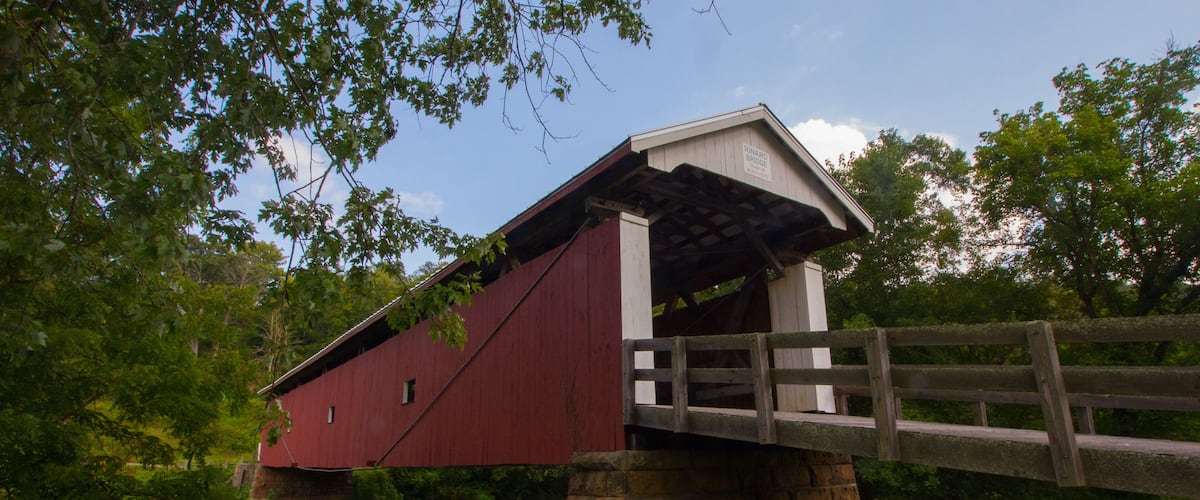 A red covered bridge with a stone foundation, surrounded by greenery and trees.