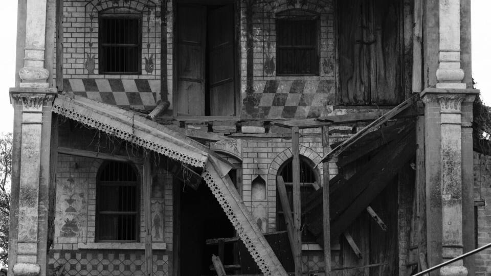 A large, ornate building with intricate architectural details, including a balcony with latticework and a staircase leading to the upper floor.