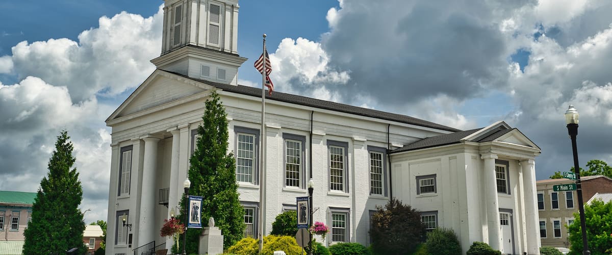 A white historic building with a clock tower, surrounded by lush greenery and a partly cloudy sky.