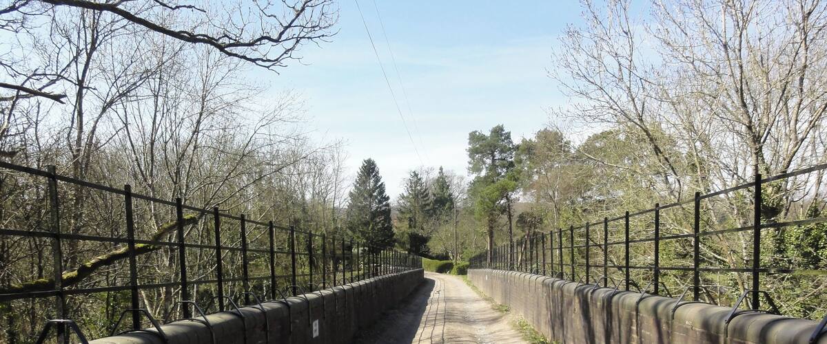 A paved pathway flanked by metal fences and brick walls, leading through a wooded area with bare trees.
