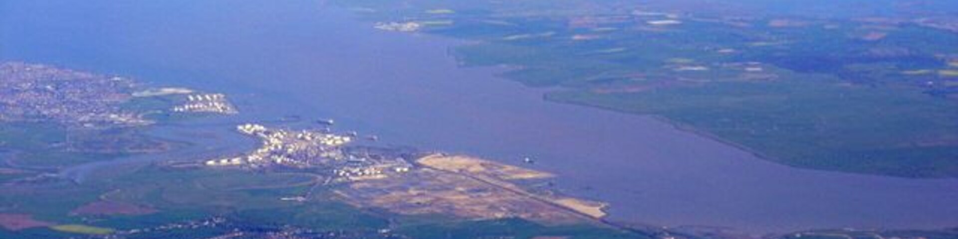 Vista aérea de uma cidade costeira com uma grande extensão de água, campos verdes e uma área urbana densa.