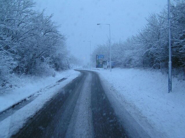 A snow-covered road flanked by trees with a streetlight in the distance.