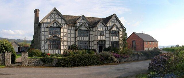 A large Tudor-style house with a stone chimney, surrounded by lush gardens and a stone wall.