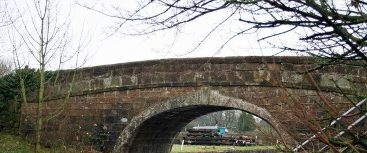 A stone bridge over a calm river with a grassy bank and bare trees.