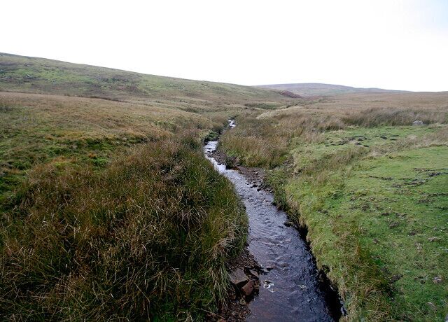 A narrow stream flowing through a grassy landscape with rolling hills in the background.