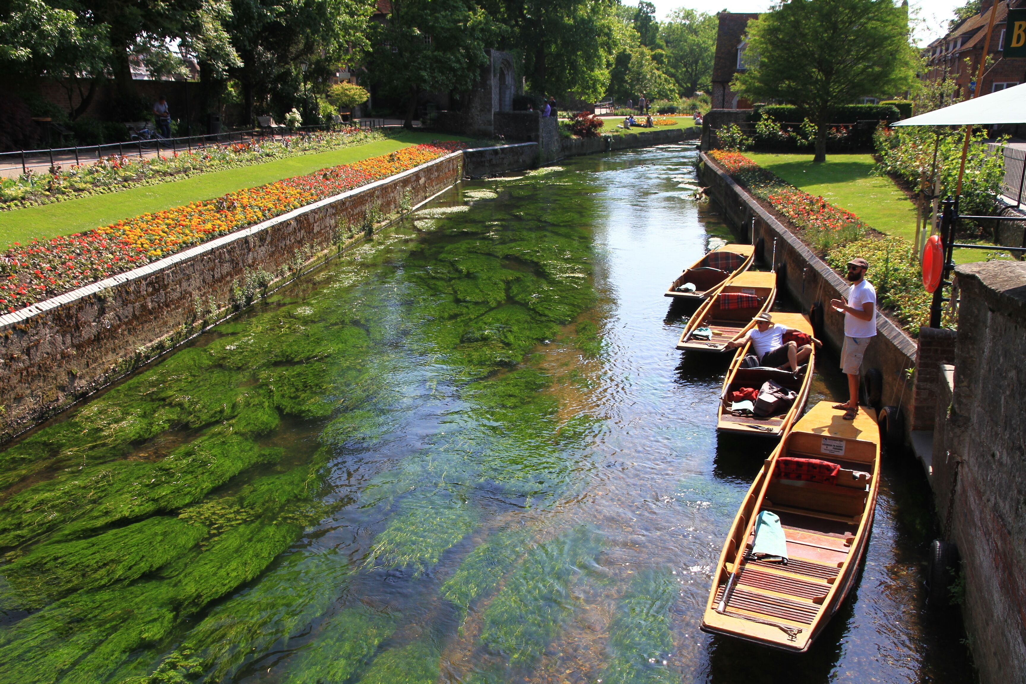 A canal with boats, greenery, and a stone wall.