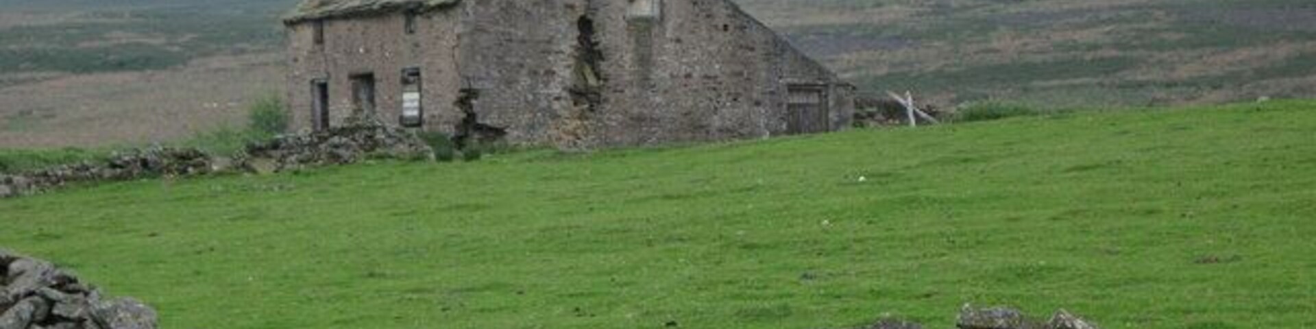 A dilapidated stone building in a grassy field with a pile of stones nearby and hills in the background.