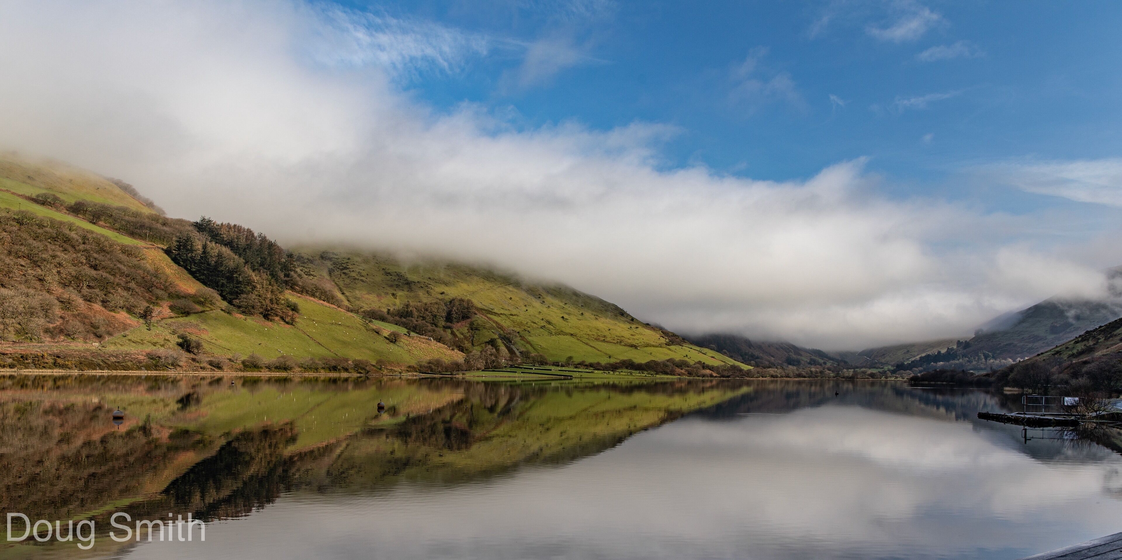 A calm lake reflecting the surrounding hills and clouds.