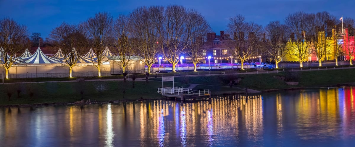 Vista nocturna de un parque con un lago, tiendas de campaña y un edificio parecido a un castillo iluminado con luces de colores.
