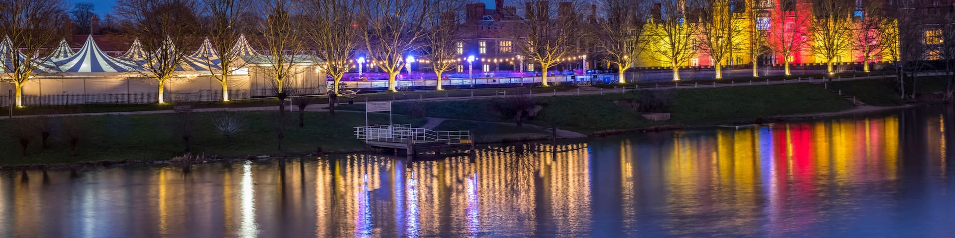A nighttime view of a park with a lake, tents, and a castle-like building illuminated with colorful lights.