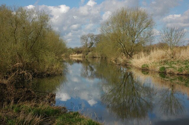 A calm river with reflections of trees and sky, surrounded by grassy banks.