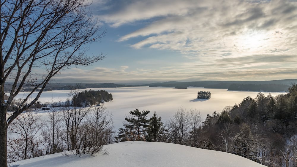 Eine verschneite Landschaft mit einem See, Bäumen und wolkenlosem Himmel.