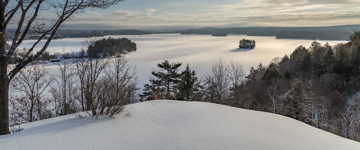 A snow-covered landscape with a lake, trees, and a clear sky.