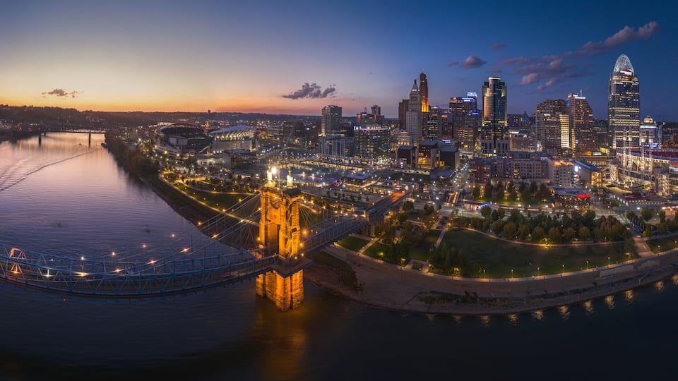 A cityscape at dusk with illuminated buildings, a river, and a bridge.