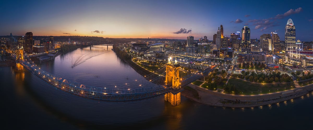 A cityscape at dusk with illuminated buildings, a river, and a bridge.