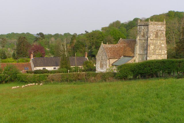 A stone church with a bell tower, surrounded by a green field and a few houses.