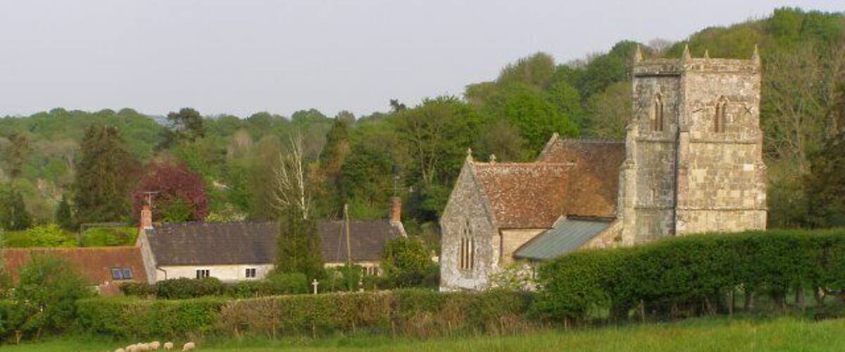A stone church with a bell tower, surrounded by a green field and a few houses.